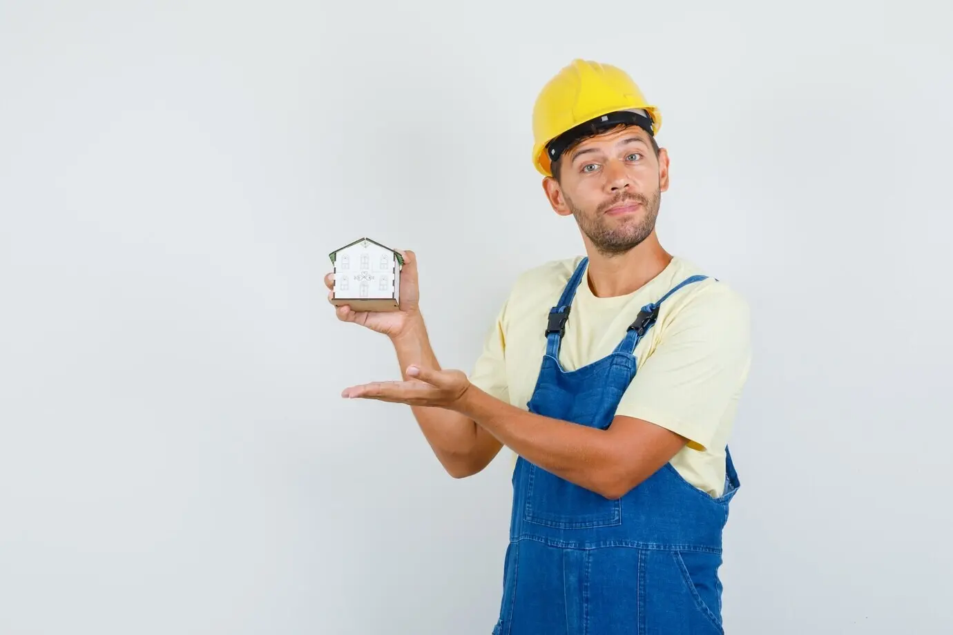 Front view of a young engineer in uniform presenting a house model with a careful expression.