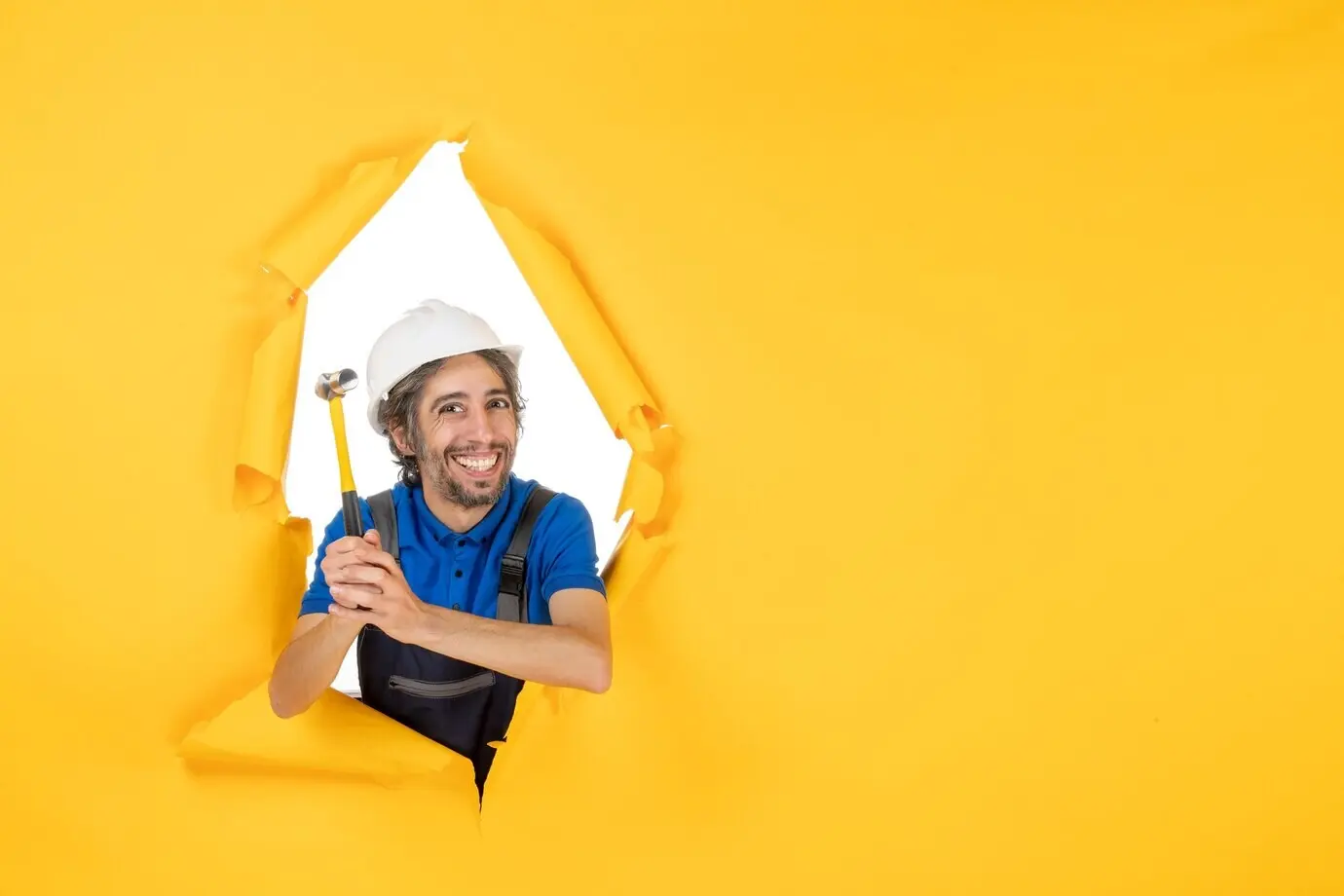 Front view of a male builder in uniform with a hammer in front of a yellow wall; worker, man, building job, constructor, architecture, color.
